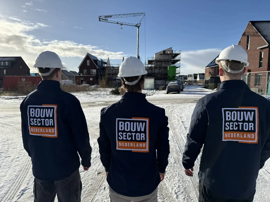 Drie vakmensen van Bouwsector Nederland met witte helm kijken uit over een nieuwbouwwijk in aanbouw
